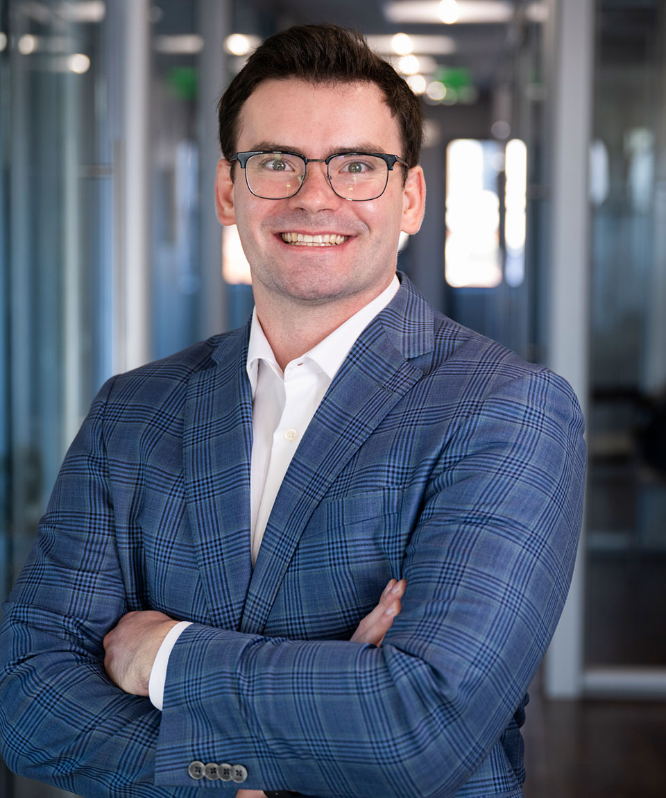 A man wearing a blue plaid sport coat, white shirt, and black glasses on his face smiles in front of a blurred office corridor.